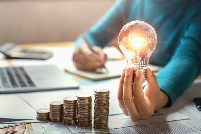 Woman hand holding lightbulb with coins stack - ZenQ Designs